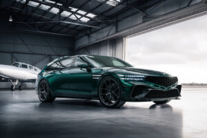 넓은 비행기 격납고 안에 세련된 짙은 녹색 스포츠 왜건이 주차되어 있고, 커다란 열린 격납고 문 근처에는 흰색 개인 제트기가 배경으로 보입니다.