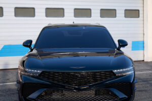 검은색 제네시스 차량이 위에 제네시스 로고가 표시된 차고 문 앞에 주차되어 있습니다. 차량이 정면을 향하고 있어 세련되고 현대적인 전면 디자인이 돋보입니다.