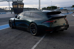 부분적으로 흐린 하늘 아래 폴 리카르 서킷의 피트 레인 근처 레이스 트랙에 한쪽 문이 위로 열린 세련된 짙은 녹색의 고급 스포츠카 한 대가 주차되어 있습니다.