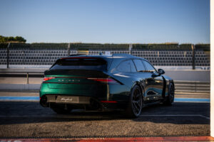 짙은 녹색 포르쉐 스포츠 왜건이 경기장 좌석과 맑고 푸른 하늘을 배경으로 피트 레인 근처의 레이싱 트랙에 주차되어 있습니다. 차량은 후방에서 낮은 각도로 보입니다.