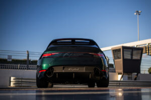 짙은 녹색의 스포츠카가 피트 레인 근처의 레이싱 트랙에 주차되어 있고, 뒤쪽에서 보면 반짝이는 표면이 보입니다. 하늘은 구름 몇 개로 맑고 경기장 조명이 배경으로 보입니다.