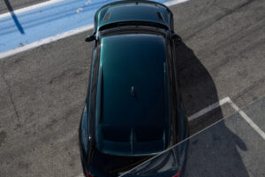 아스팔트와 파란색 레이스 트랙 라인으로 둘러싸인 피트 레인에 주차된 짙은 녹색 스포츠카의 오버헤드 뷰입니다. 펜스의 그림자가 장면을 부분적으로 가로지릅니다.