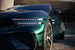 야외에 주차된 반짝이는 녹색 스포츠카를 클로즈업하여 전면 왼쪽 헤드라이트, 휠, 날렵한 차체 라인에 초점을 맞추고 배경에 흐릿한 날개 로고가 보이는 장면입니다.