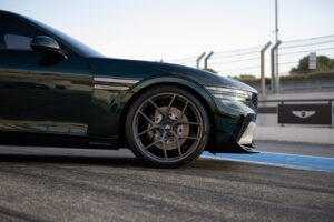 레이싱 트랙에 주차된 짙은 녹색의 고급 스포츠카를 클로즈업하여 앞바퀴, 날렵한 차체 라인, 독특한 합금 림을 보여주고 금속 울타리와 표지판을 배경으로 보여줍니다.