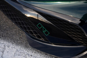 녹색으로 윤곽이 그려진 기하학적 로고가 있는 짙은 녹색 자동차의 전면 하단 범퍼를 클로즈업합니다. 흰색과 파란색으로 칠해진 선이 있는 포장 도로 위에 자동차가 주차되어 있습니다.