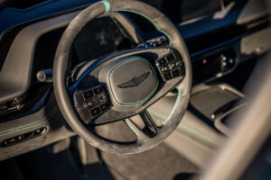 녹색 스티치 디테일이 있는 스티어링 휠과 중앙의 날개 로고에 초점을 맞춘 자동차 내부 클로즈업, 열린 운전석 도어를 통해 햇빛이 비추는 모습입니다.